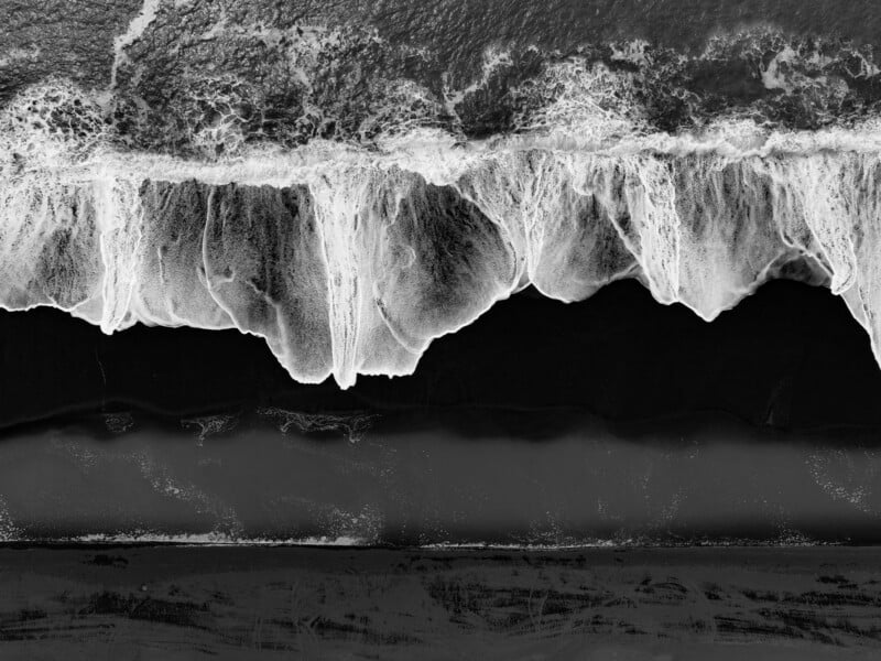 An aerial view shows white ocean waves crashing onto a dark, black sand beach.