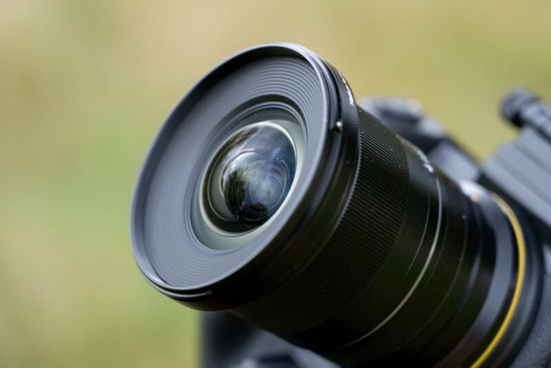 A close-up view of a camera lens with a prominent, rounded front element against a soft, out-of-focus green background.