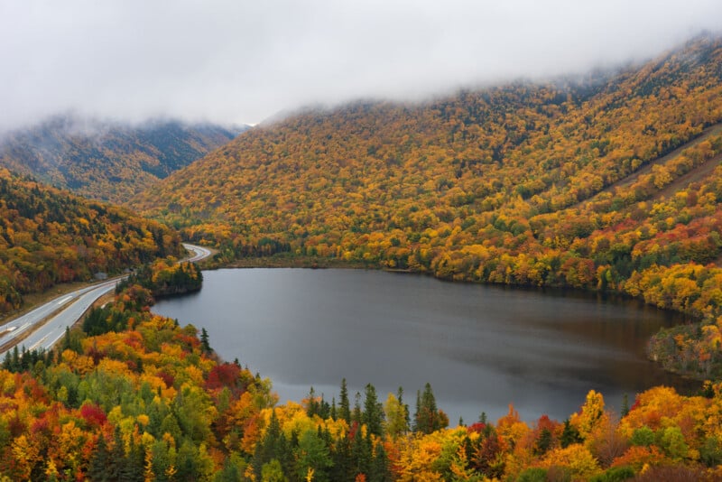 A highway curves along the edge of a dark lake surrounded by dense, colorful autumn forests under a cloudy, mountainous sky.