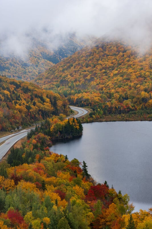 A winding road curves along the shore of a lake surrounded by dense, autumn-colored forests under a cloudy, misty sky.