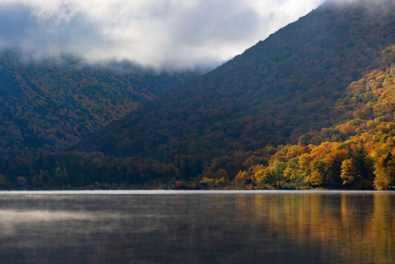A calm lake reflects a mountain slope covered in dense, colorful autumn foliage under a cloudy sky with low-hanging mist.