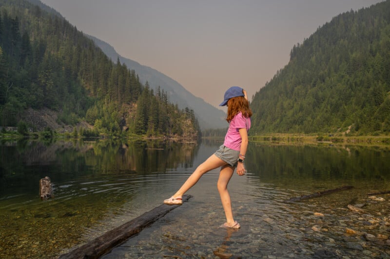 A girl in a pink shirt and blue cap balances on a log in a shallow, clear lake surrounded by forested mountains under a hazy sky.