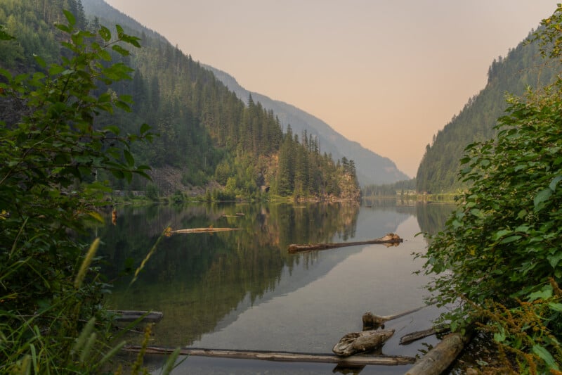A calm lake reflects the surrounding evergreen mountains under a hazy, smoke-filled sky, with several logs floating in the foreground.