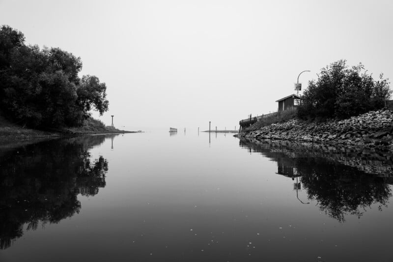A calm, reflective body of water flows between a tree-lined bank on the left and a rocky shore with a small building on the right.