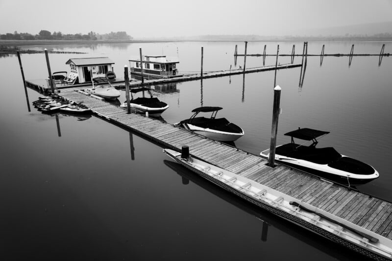 A wooden dock extends into calm water, featuring a small building, a houseboat, and several motorboats and jet skis moored alongside.