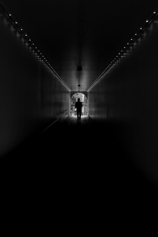 A person walks away through a dark, narrow tunnel illuminated by two rows of overhead lights, heading toward a bright exit.