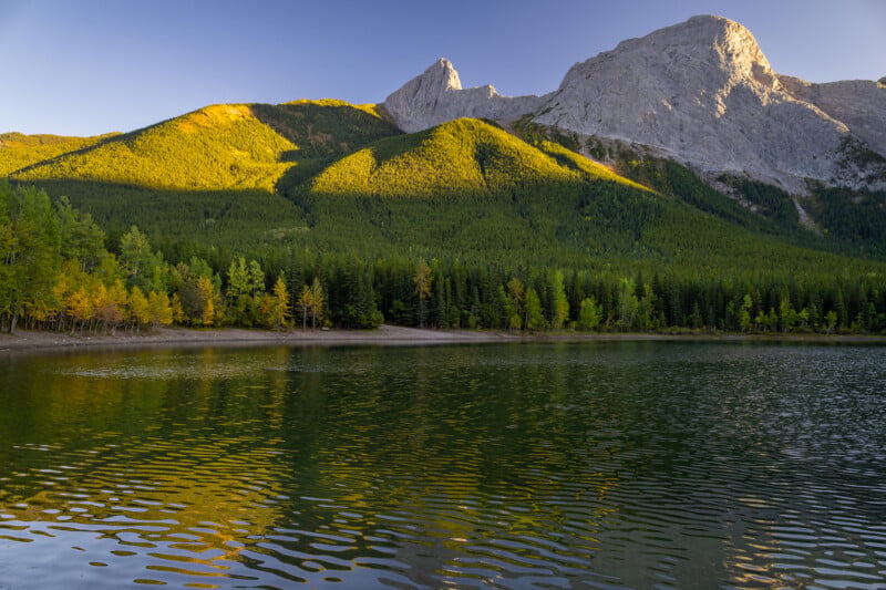 A calm lake reflects the golden sunlight hitting a forested mountain slope beneath a jagged, rocky peak.