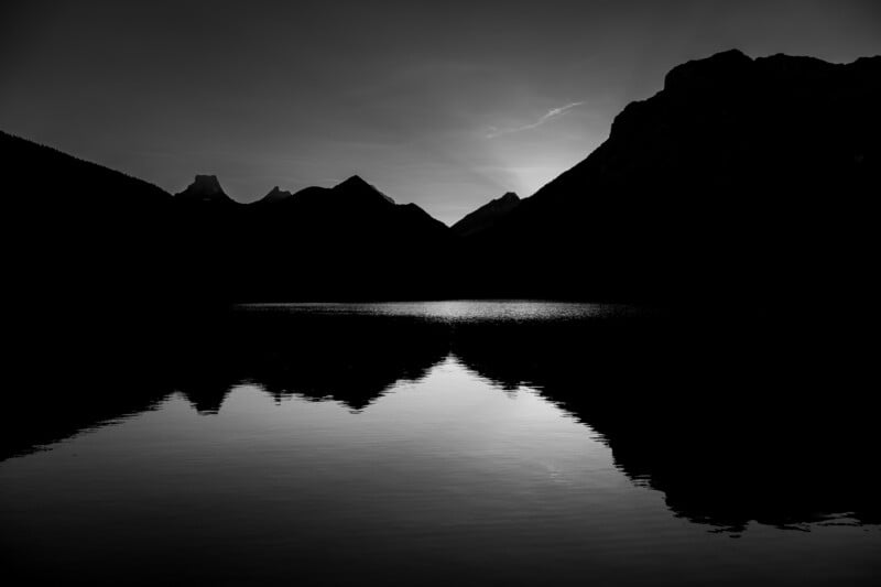 A black and white silhouette of mountain peaks reflected in the calm, dark water of a lake at sunset.