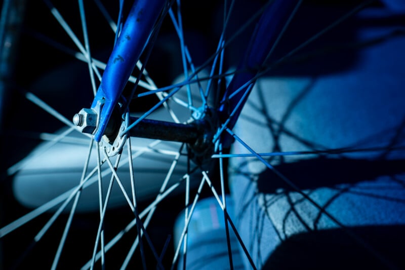A close-up view of a blue bicycle fork and front wheel hub with metal spokes, illuminated by cool blue light.