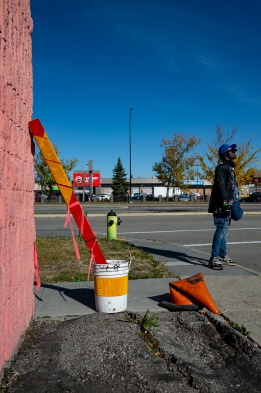 A person wearing a blue cap and dark clothing stands on a sidewalk near a pink wall, a construction marker in a bucket, and a flattened orange traffic cone.