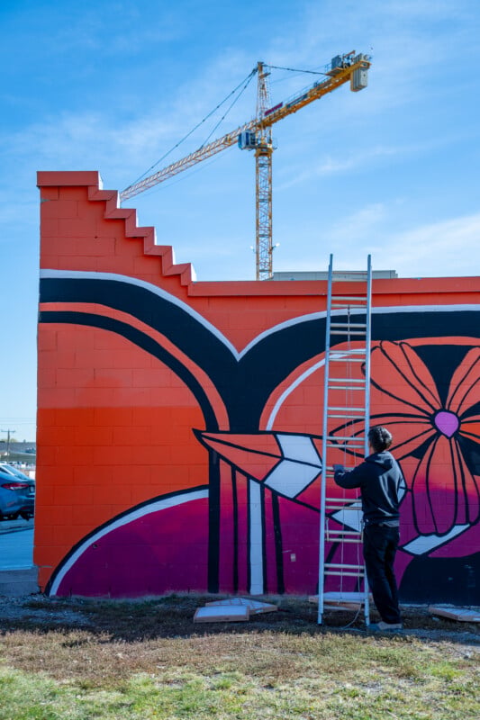 A person in a black hoodie stands on a ladder painting a large orange and black mural on a brick wall, with a construction crane visible in the background.