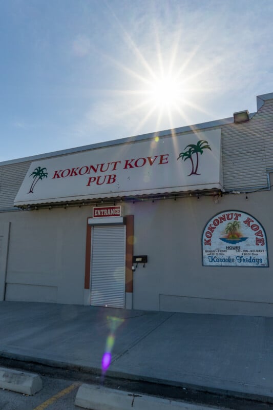 The exterior of the Kokonut Kove Pub, featuring a closed metal roll-down door, a sign with operating hours, and a bright sunburst in the sky above.