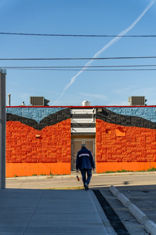A person wearing a blue jacket walks toward a building with a textured wall painted in horizontal bands of orange, black, and light blue.