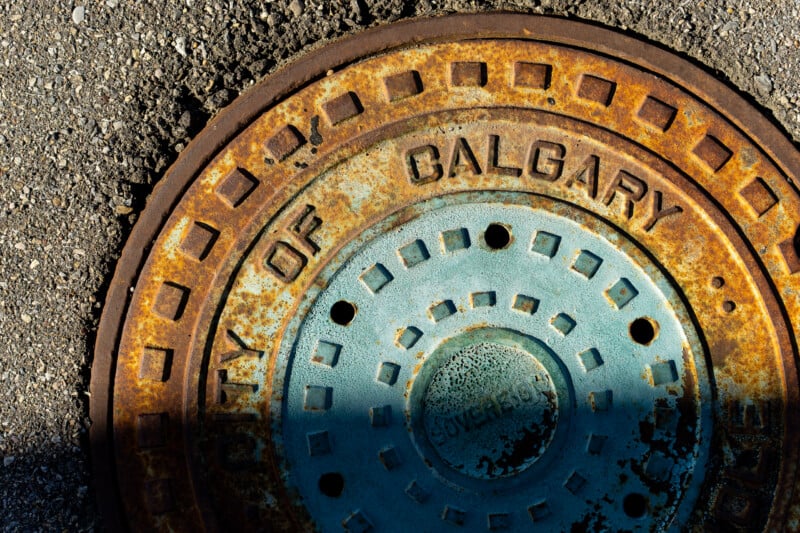 A close-up, high-angle shot of a weathered, circular metal manhole cover labeled "CITY OF CALGARY" set into asphalt.