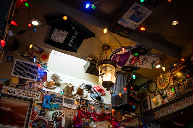 A cluttered ceiling and wall in a pub are decorated with string lights, hanging t-shirts, cowboy hats, vintage posters, license plates, and a central lantern.