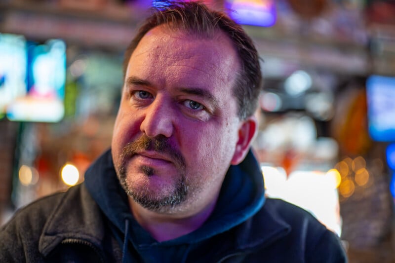 A middle-aged man with a mustache and goatee looks directly at the camera in a dimly lit, blurred setting with colorful ambient lighting.