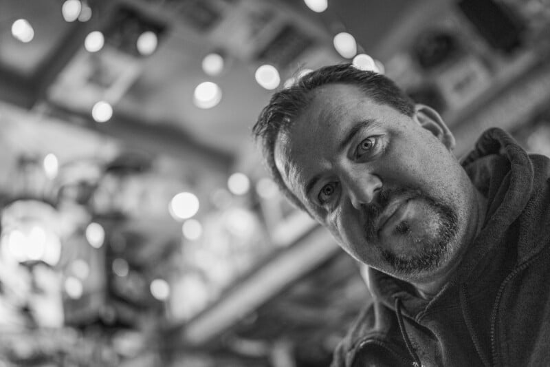 A black-and-white close-up portrait of a man with a goatee, looking directly at the camera against a blurred, bokeh background.