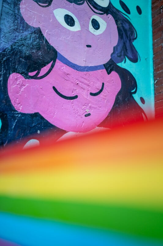 A mural of two stylized faces, one with open eyes and one with closed eyes, is partially obscured by a blurred rainbow flag.