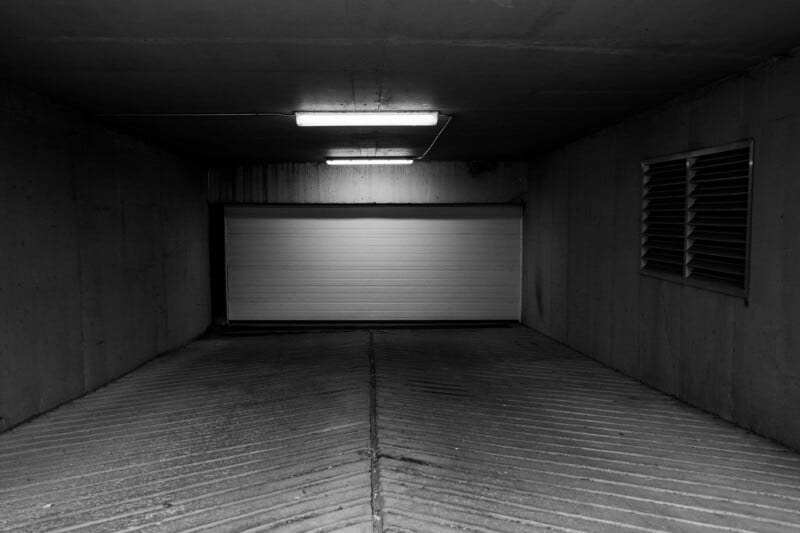 A black and white view of a concrete garage entrance with a closed white door, illuminated by two overhead fluorescent lights.