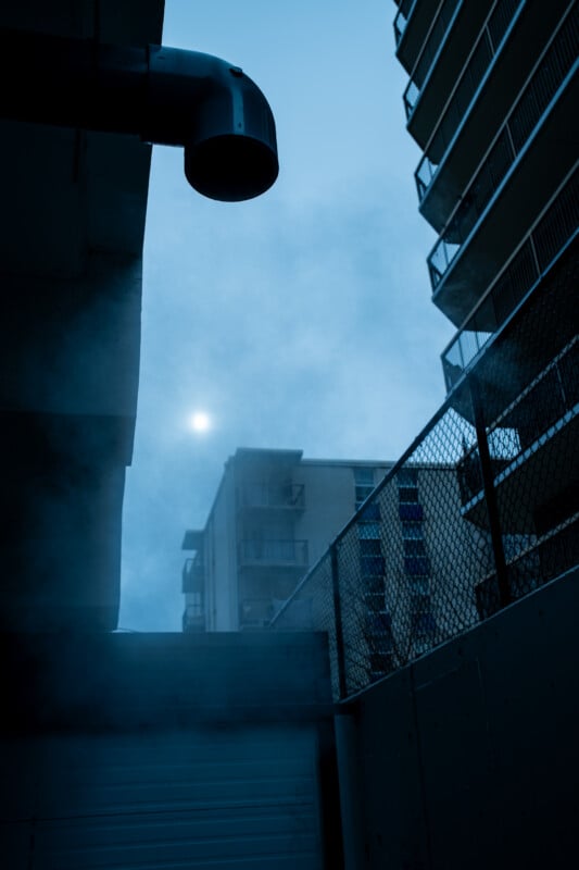 A low-angle view of a dark, foggy urban alleyway with a large drainage pipe overhead and apartment buildings in the distance.