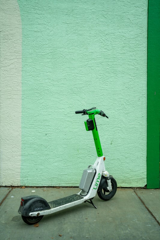 A white and green Lime electric scooter stands on a concrete sidewalk against a textured light green wall.
