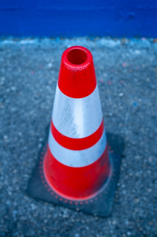 A traffic cone with orange and white reflective stripes stands on a gray asphalt surface in front of a blue wall.