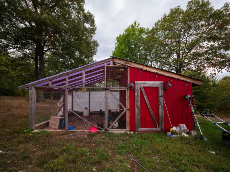A red wooden chicken coop with a wire-fenced run sits on a grassy lawn under a cloudy sky, with trees in the background.