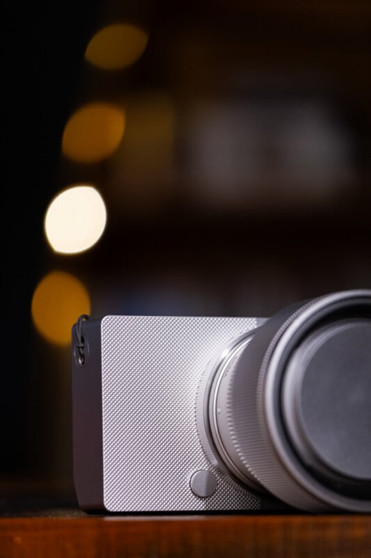 A silver camera with a textured front panel sits on a wooden surface against a dark background with blurred, circular golden lights.
