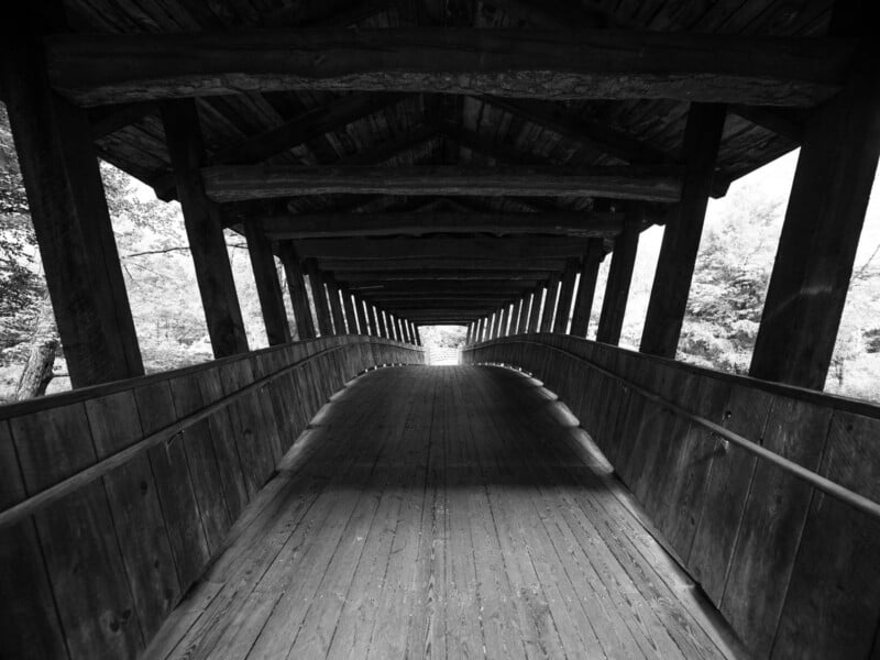 A black-and-white, eye-level view looking through the interior of a wooden covered bridge toward the light at the far end.
