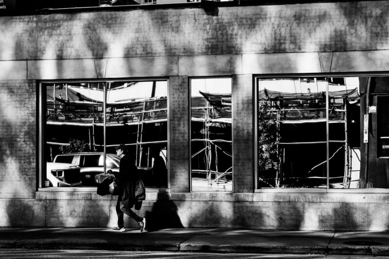 A person in a cap and dark clothing walks along a sidewalk past a building with large windows reflecting construction scaffolding.