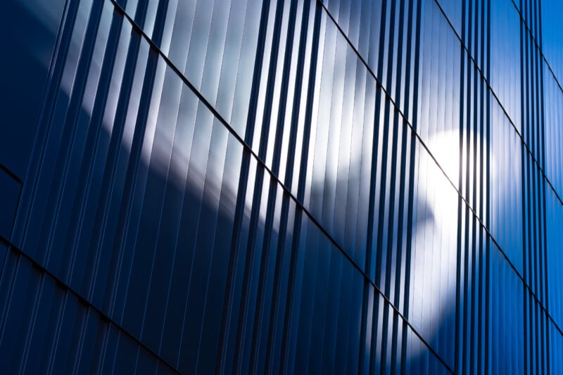 A close-up view of a modern building facade featuring vertical metal panels with a bright sun reflection and cloud patterns visible on the surface.