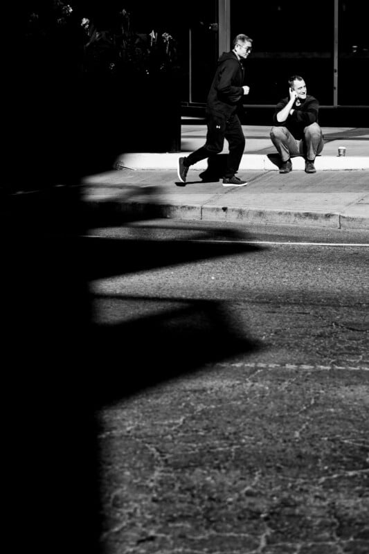 A man in a hoodie walks past another man sitting on a curb and talking on a cell phone in a sunlit urban area.