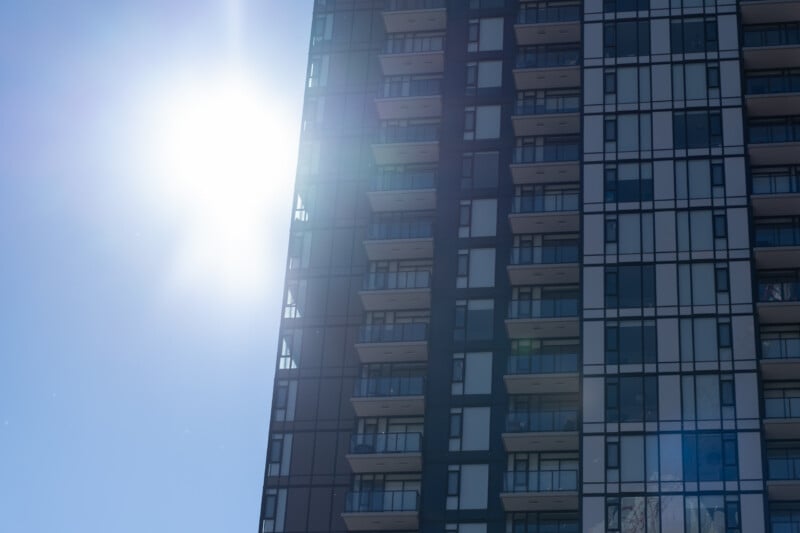 A modern high-rise apartment building with glass balconies and large windows is silhouetted against a bright, sunlit sky.
