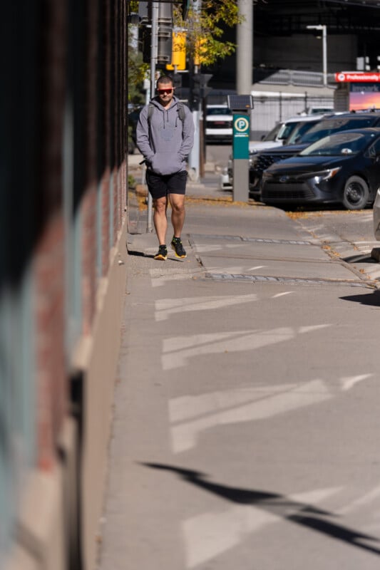 A man in a grey hoodie, black shorts, and sunglasses walks along a sidewalk next to a brick building.
