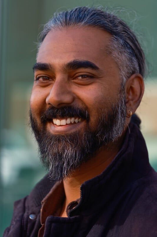 A close-up portrait of a smiling man with a salt-and-pepper beard, long graying hair pulled back, and a small gold hoop earring in his left ear.