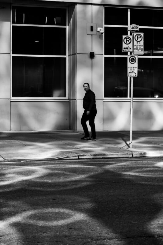 A man in dark clothing walks along a sidewalk past a building with large windows and several parking regulation signs.