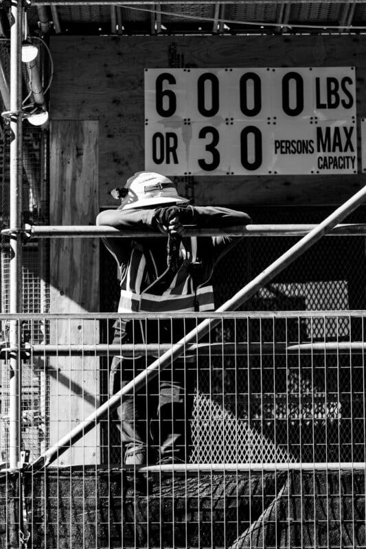 A construction worker in a hard hat and high-visibility vest leans against a metal railing in front of a capacity sign.