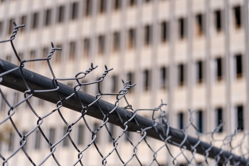A close-up view of a chain-link fence with twisted wire tops, set against a blurred background of a tall office building.