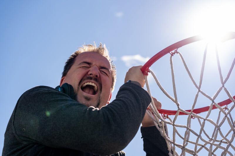 A man with his eyes closed and mouth open in an expression of intense emotion reaches up to grab a basketball hoop.