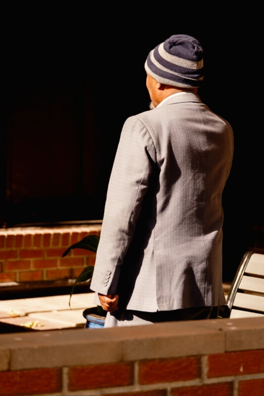 A person wearing a grey suit and a striped beanie stands with their back to the camera near a brick wall.