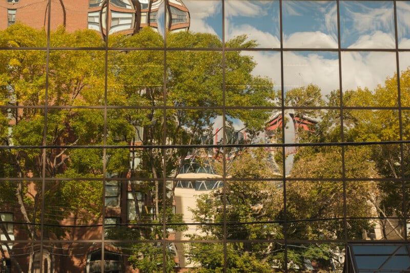 A grid of glass windows reflects a scene of green and yellow autumn trees, a brick building, and a blue sky with white clouds.