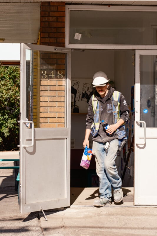 A construction worker in a hard hat and safety vest walks out of a building doorway at address 348, carrying a bag of chips and a water bottle.
