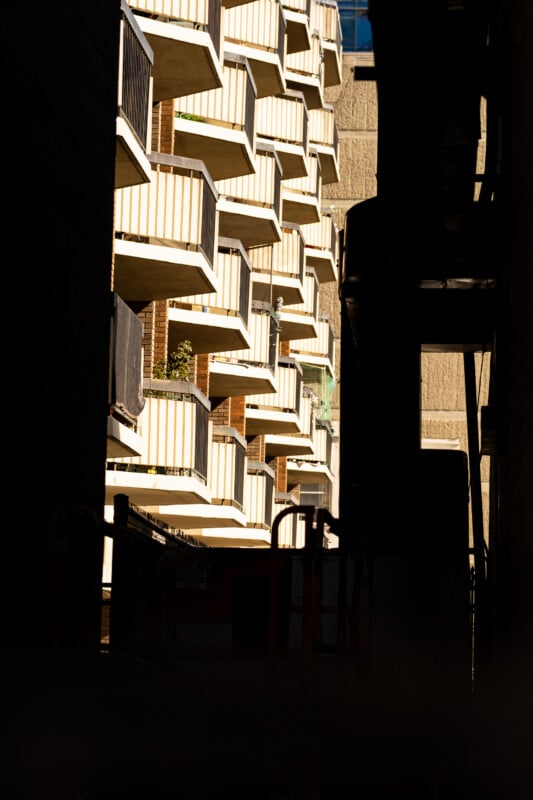 A view through a dark, narrow alleyway looking toward a brightly lit apartment building with stacked balconies.
