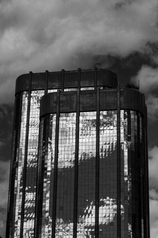A black and white low-angle view of a modern skyscraper with a glass facade reflecting cloudy skies.