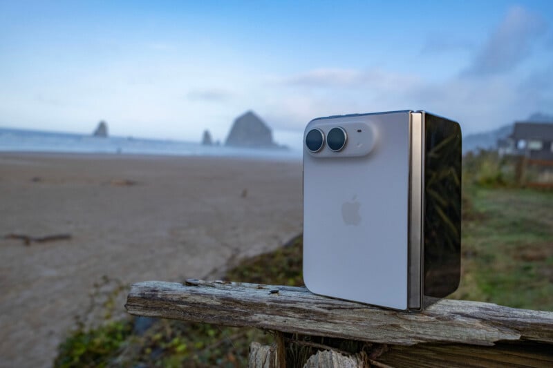 A white, foldable smartphone with a dual-lens camera stands upright on a weathered wooden post overlooking a beach.