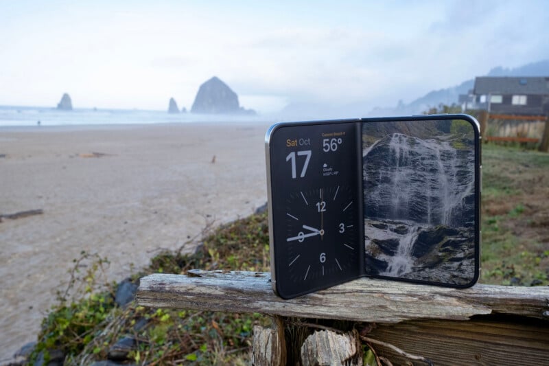 A foldable smartphone sits open on a weathered wooden fence post at a beach, displaying a clock and a waterfall image, with Haystack Rock in the background.