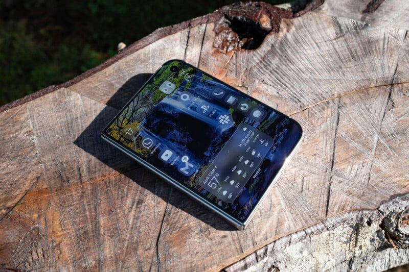 A smartphone with a waterfall wallpaper and app icons sits on a textured wooden tree stump.