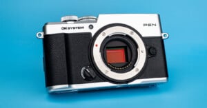 A silver and black OM SYSTEM PEN mirrorless camera body sits on a bright blue background, showing the lens mount and sensor.