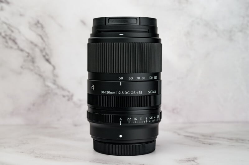 A black Sigma 50-120mm f/2.8 DC OS lens stands vertically against a white marble background.