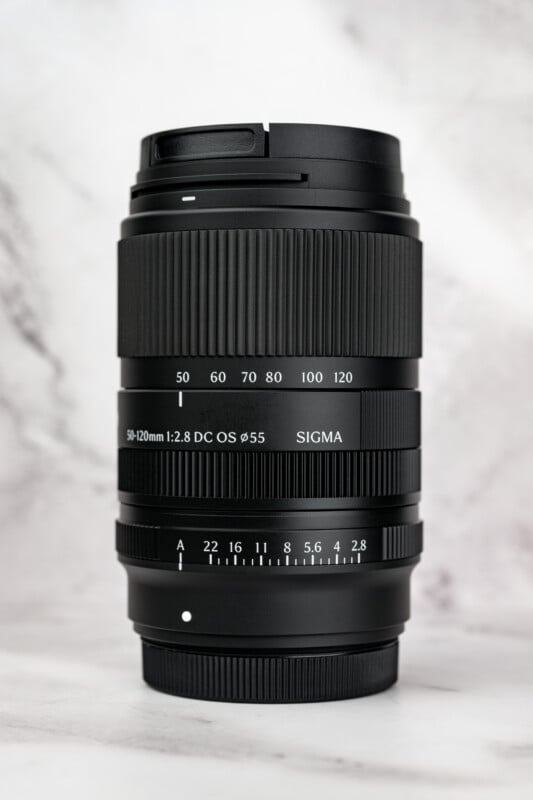 A black Sigma 50-120mm f/2.8 camera lens standing vertically against a white marble background.